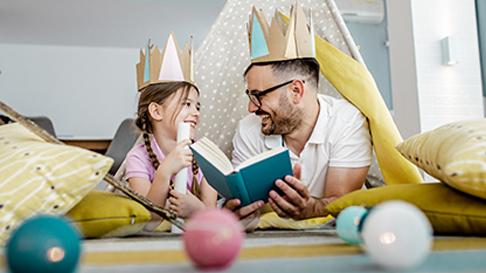 Father and daughter wearing crowns reading a book and smiling.